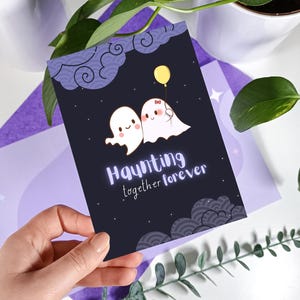 May include: A black card with a white ghost couple holding a yellow balloon. The card has a purple and white background with a repeating pattern of clouds. The text on the card reads "Haunting together forever."