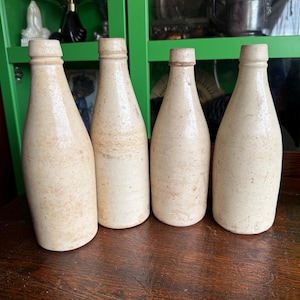 May include: Four vintage stoneware bottles with a light brown glaze. The bottles are all the same size and shape, and they are arranged in a row on a wooden surface.