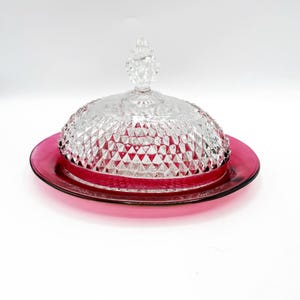 May include: A clear glass butter dish with a domed lid and a diamond pattern. The dish sits on a red glass base.
