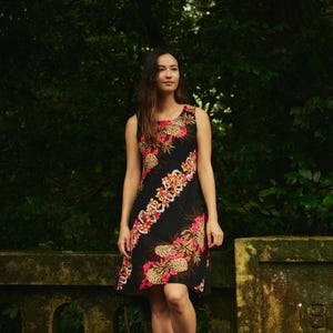 May include: A sleeveless black dress with a floral pattern featuring pink, yellow, and orange flowers. The dress has a scoop neckline and a diagonal floral design across the front. The model is standing outdoors.