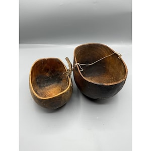 May include: Two wooden bowls with a rustic appearance. The bowls are different sizes and have a dark brown color. One bowl has a small rope attached. The bowls appear to be handmade and have a natural wood grain.