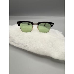 May include: Black and silver-toned sunglasses with green lenses. The sunglasses have a classic design with a black upper frame and a silver-toned lower frame. The lenses are a light green color.