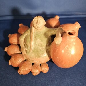 May include: A terracotta-colored ceramic sculpture depicting a figure in a green garment holding a large pot. The sculpture has several smaller pots around the figure. The pot held by the figure has the inscription "Amy 92".