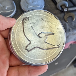 May include: A silver metal coaster with a stylized bird design. The bird is in flight and has a long beak. The coaster is round and has a raised edge.