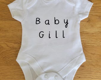 May include: White baby onesie with the text "Baby Gill" in black. The onesie has short sleeves and snaps at the bottom. The garment is made of soft material and is designed for a baby.