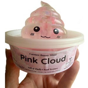 Puede incluir: Un recipiente de slime rosa etiquetado como "Pink Cloud" con una cara de dibujos animados en la parte superior. El slime tiene una textura de nube suave y esponjosa. El recipiente es blanco con una etiqueta rosa y el texto "Carmee Bunny Slime".
