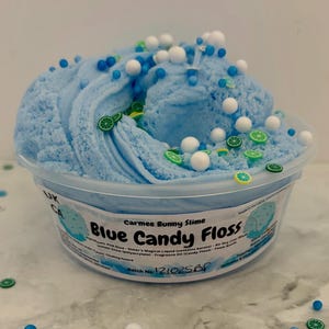 Peut inclure: Un pot de slime bleu, appelé "Blue Candy Floss", orné de sphères blanches et bleues, et de décorations en forme de citron vert. Le récipient porte l'étiquette "Carmee Bunny Slime". Le produit est présenté dans un récipient en plastique transparent.