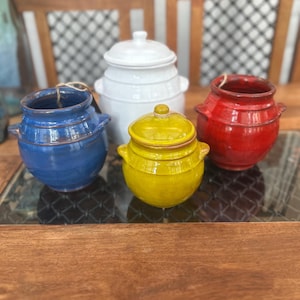 May include: Four brightly coloured ceramic jars with lids. The jars are in blue, white, yellow, and red. Each jar has a rounded shape with small handles. The jars are displayed on a glass surface.
