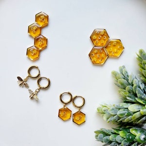 May include: Gold tone earrings with a honeycomb design and a bee charm. The honeycomb earrings are filled with small yellow flowers. The bee charm is attached to a hoop earring.