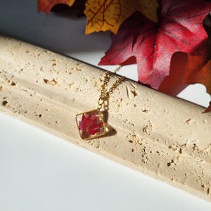 May include: A gold-colored chain necklace with a square pendant. The pendant contains a red maple leaf encased in clear resin. The background includes a textured, light-colored surface and red and yellow autumn leaves.