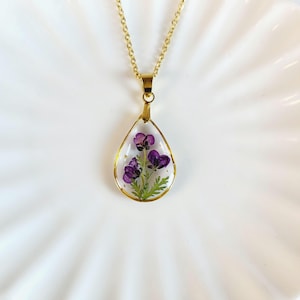 May include: A gold chain necklace with a teardrop-shaped pendant featuring a pressed purple flower inside.
