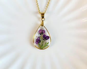 May include: A gold-toned teardrop pendant necklace featuring a clear resin center that encapsulates small, pressed purple flowers with green stems and leaves. The pendant hangs from a delicate gold-toned metal chain with a polished finish.