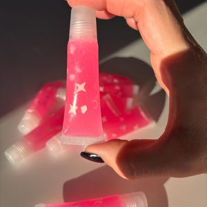 May include: A bright pink lip gloss tube with white star and dot designs. The tube is held up by a hand with a black painted nail. Several other tubes are visible in the background. The gloss is in a clear tube.