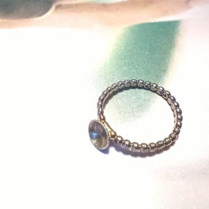 May include: A silver ring with a beaded band and a central, oval-shaped, iridescent stone. The ring's design is simple, with the stone set in a gold-colored bezel. The ring is likely made of sterling silver.