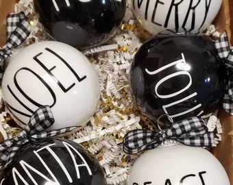 May include: Six black and white Christmas ornaments with black and white buffalo plaid ribbon. The ornaments have the words "Hope", "Merry", "Noel", "Jolly", "Santa", and "Peace" written in black on a white background or white on a black background.