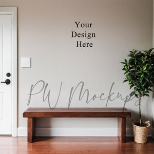 Puede incluir: Un banco de madera con un acabado marrón claro se encuentra frente a una pared beige. El banco tiene un diseño sencillo con líneas rectas y una forma rectangular. Una pequeña planta en maceta con hojas verdes se encuentra a la derecha del banco. La pared tiene el texto "Your Design Here" escrito en ella.