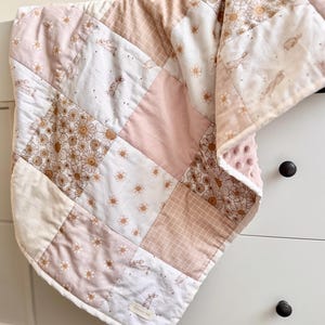 May include: A patchwork quilt in shades of pink, white, and tan. The quilt features floral, bunny, and grid patterns. The quilt is draped over a white chest of drawers. The quilt is a baby blanket.
