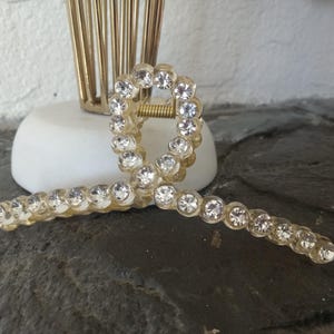 May include: A gold hair clip with a clear plastic base and a row of clear rhinestones around the edge. The clip is shaped like a bow.