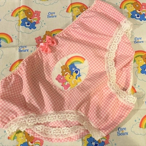 May include: Pink gingham-patterned garment with white lace trim and a pink bow. A circular patch features Care Bears under a rainbow. The background is a Care Bears print with rainbows and bears.