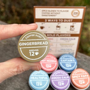 May include: A round, olive-green Coffee Dust spice blend container labeled Gingerbread, which serves 12. Other colorful containers labeled Hot Momma, Captain's, Healthful, Cocoa Calm, and Spicy Papa are visible, along with an informational card.