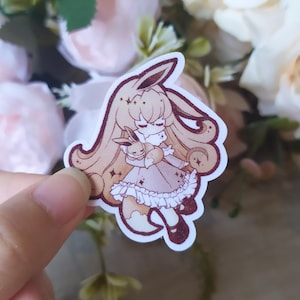 May include: A sticker featuring a cartoon illustration of a light brown-haired girl with a fox-like tail and ears, wearing a pink dress and holding a small brown fox in her arms. The girl has a light brown dress with a white lace trim and a pink bow. The sticker has a white background.