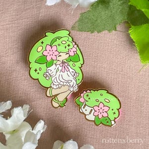 May include: Two enamel pins featuring a green-haired character with pink flower accents. The character is wearing a white dress and has a gold outline. One pin is a full figure, the other is a headshot. The pins are on a pink background.