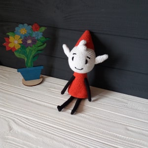May include: A crocheted elf doll with a red hat and a white body. The elf is sitting on a white wooden surface. The elf has black eyes, a black mouth, and a black nose. The elf has black arms and legs.