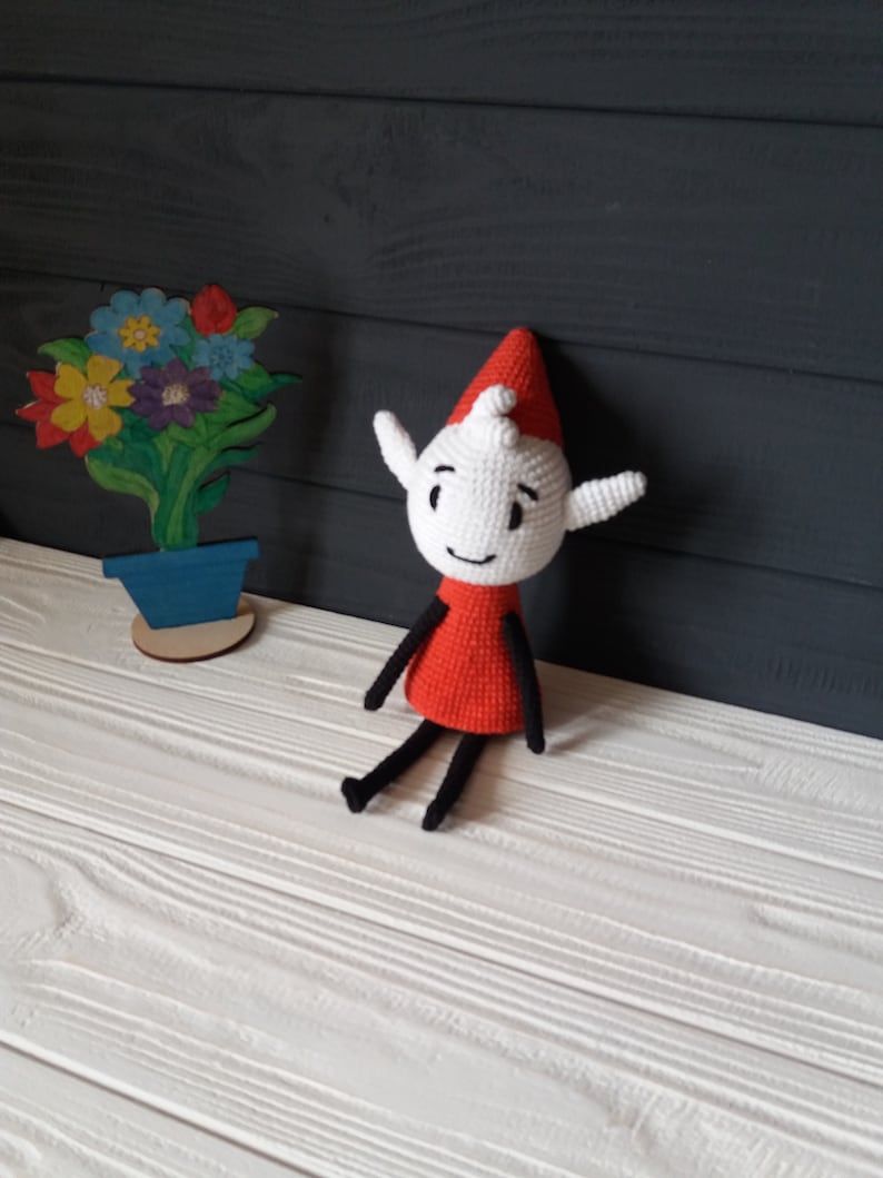 May include: A crocheted elf doll with a red hat and a white body. The elf is sitting on a white wooden surface. The elf has black eyes, a black mouth, and a black nose. The elf has black arms and legs.