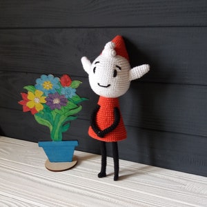 May include: A crocheted elf doll with a red dress and black legs stands next to a painted wooden flower pot with blue paint. The elf has a white face with black eyes and a smiling mouth.