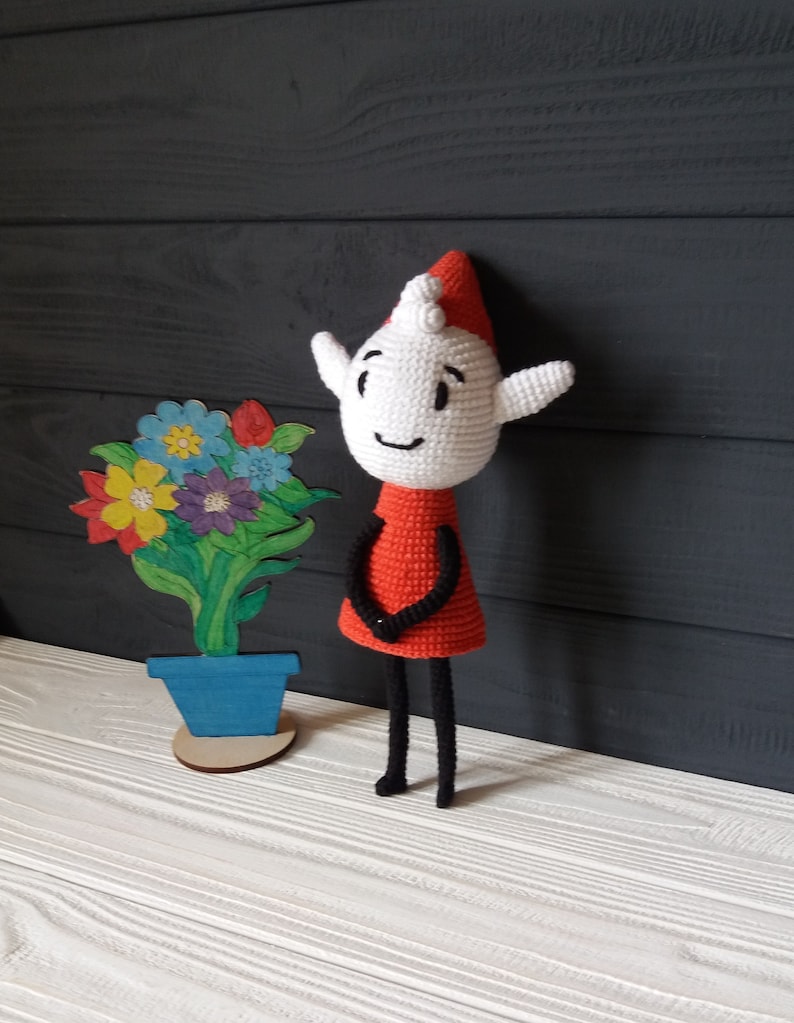 May include: A crocheted elf doll with a red dress and black legs stands next to a painted wooden flower pot with blue paint. The elf has a white face with black eyes and a smiling mouth.