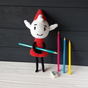 May include: A crocheted elf doll wearing a red dress and a white hat holds a blue pencil. The doll is standing on a white surface with four colored pencils and a white eraser.