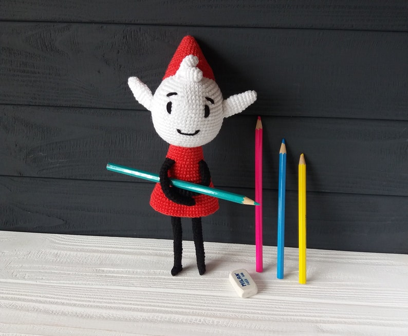 May include: A crocheted elf doll wearing a red dress and a white hat holds a blue pencil. The doll is standing on a white surface with four colored pencils and a white eraser.