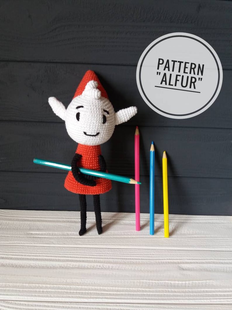 May include: Crocheted elf doll with a red dress and black legs holding a blue pencil. The doll is standing on a white wooden surface with three colored pencils behind it. The text "PATTERN "ALFUR"" is in a circle above the doll.