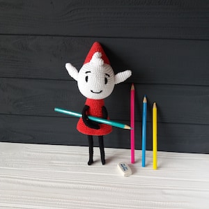 May include: A crocheted elf doll wearing a red dress and a white hat, holding a teal colored pencil. The elf is standing on a white wooden surface with four colored pencils and a white eraser.