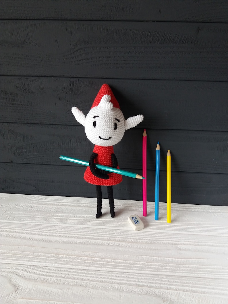May include: A crocheted elf doll wearing a red dress and a white hat, holding a teal colored pencil. The elf is standing on a white wooden surface with four colored pencils and a white eraser.