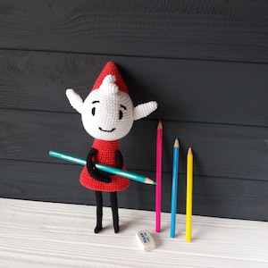 May include: A white crocheted elf doll wearing a red dress and a red hat, holding a blue pencil. The doll is standing on a white wooden surface with four colored pencils and a white eraser.