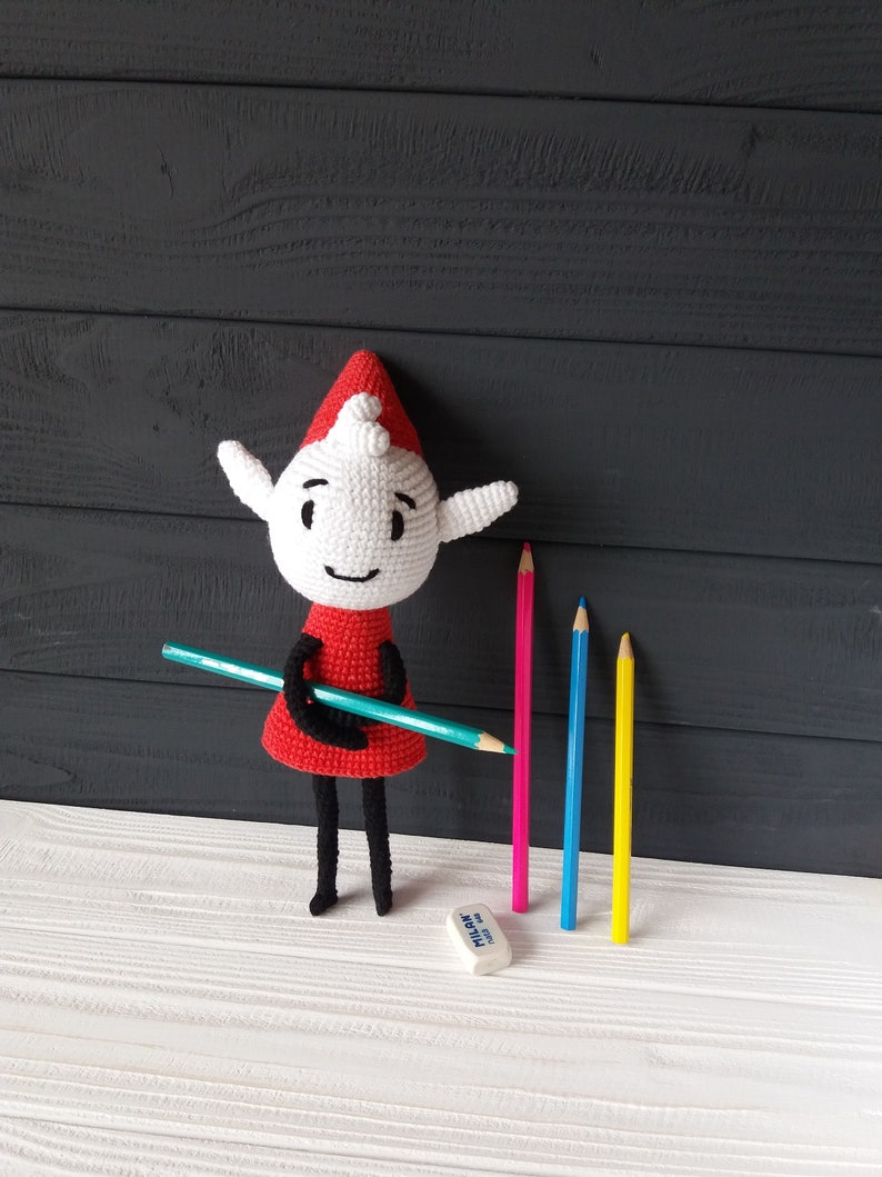May include: A white crocheted elf doll wearing a red dress and a red hat, holding a blue pencil. The doll is standing on a white wooden surface with four colored pencils and a white eraser.