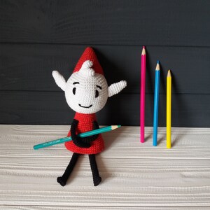 May include: A white crocheted elf doll with a red hat and a green pencil. The elf is sitting on a white wooden surface with three colored pencils behind it.
