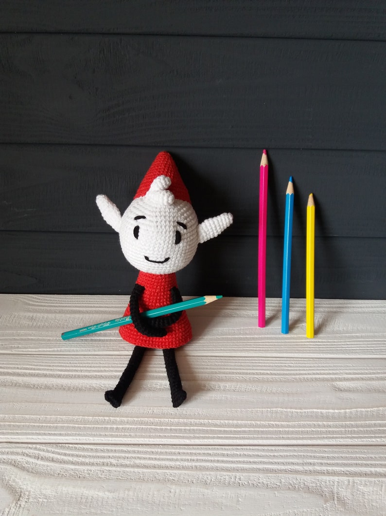May include: A white crocheted elf doll with a red hat and a green pencil. The elf is sitting on a white wooden surface with three colored pencils behind it.