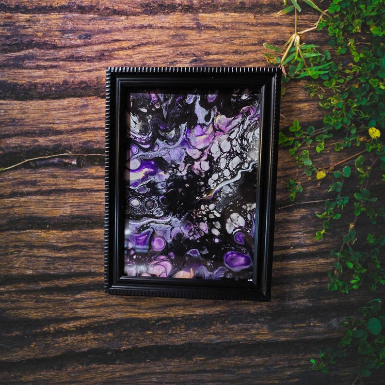 May include: A framed abstract painting with swirling purple, black, and white paint. The painting is in a black frame with a decorative border.