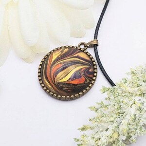 May include: A round pendant necklace with a black cord. The pendant features a swirling abstract design in shades of orange, yellow, brown, and black, encased in a bronze-colored frame.