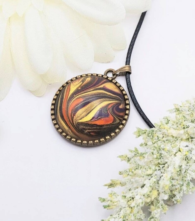 May include: A round pendant necklace with a black cord. The pendant features a swirling abstract design in shades of orange, yellow, brown, and black, encased in a bronze-colored frame.
