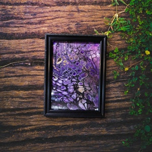 May include: A framed abstract painting with swirling purple and gold paint. The painting is in a black frame with a decorative border.