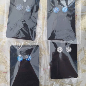 May include: Four pairs of earrings displayed on black cards. The earrings are made of silver and have round glass cabochons with different designs. The designs include a blue and white marble pattern, a blue and white swirl pattern, and a white and gray marble pattern.