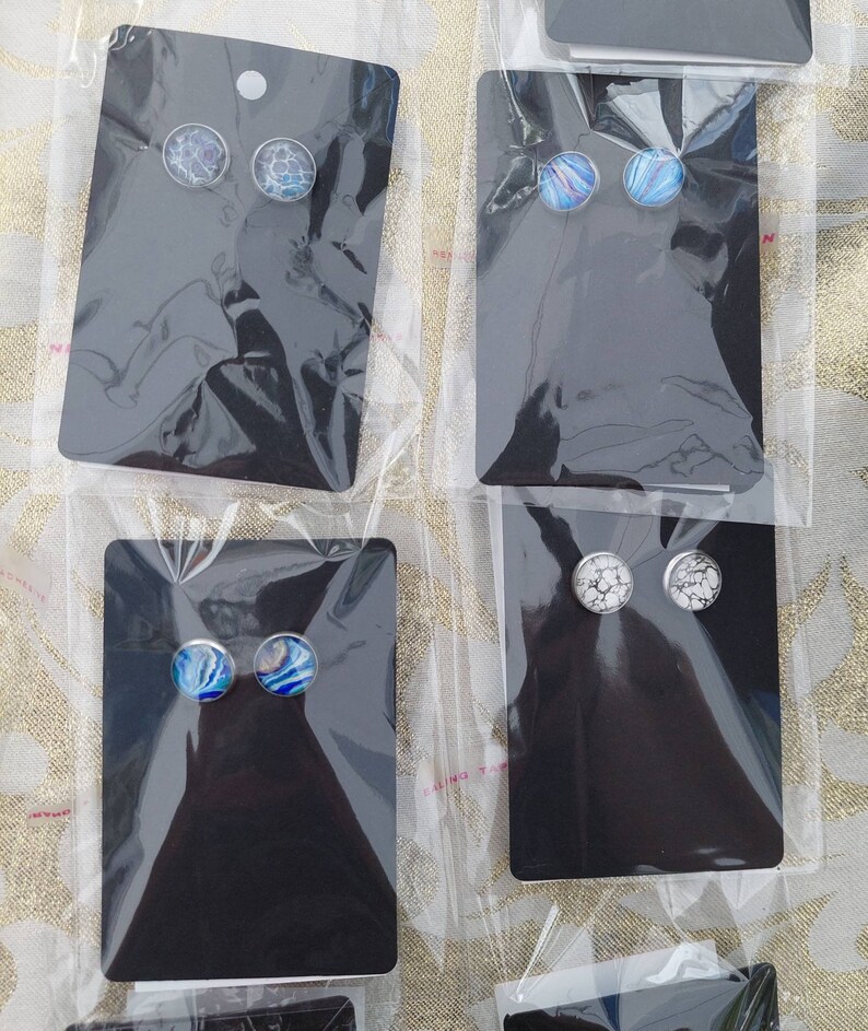 May include: Four pairs of earrings displayed on black cards. The earrings are made of silver and have round glass cabochons with different designs. The designs include a blue and white marble pattern, a blue and white swirl pattern, and a white and gray marble pattern.
