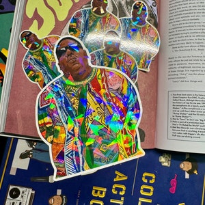 May include: A holographic sticker featuring a colorful portrait of the rapper The Notorious B.I.G. in a patterned shirt. The sticker is a vibrant mix of rainbow colors and has a glossy finish.