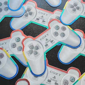 May include: A collection of gray PlayStation controller-shaped stickers. Each sticker has a colorful border and features the word "COZY" above the PlayStation logo, with "SELECT" and "START" buttons. The controllers are scattered across a dark surface.