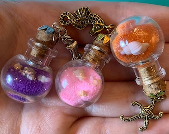 Könnte beinhalten: Drei kleine Glasflaschen mit Korkstopfen, jede gefüllt mit buntem Sand und Muscheln. Die Flaschen sind an einem goldfarbenen Seepferdchen-Anhänger und einem Seestern-Anhänger befestigt.