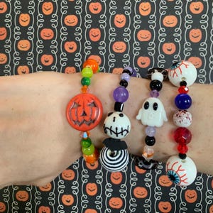 以下が含まれることがあります： オレンジ色のカボチャ、幽霊、眼球のチャームが付いたハロウィーンをテーマにしたブレスレット。ブレスレットは、さまざまな形とサイズのカラフルなビーズが特徴です。背景はカボチャのハロウィーンをテーマにしたパターンです。