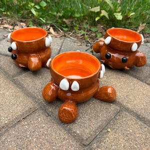 May include: Three brown ceramic bowls with orange interiors, shaped like cartoon bears. Each bowl has white accents, black eyes, and rounded feet. The bowls are displayed on a brick surface, with greenery in the background.